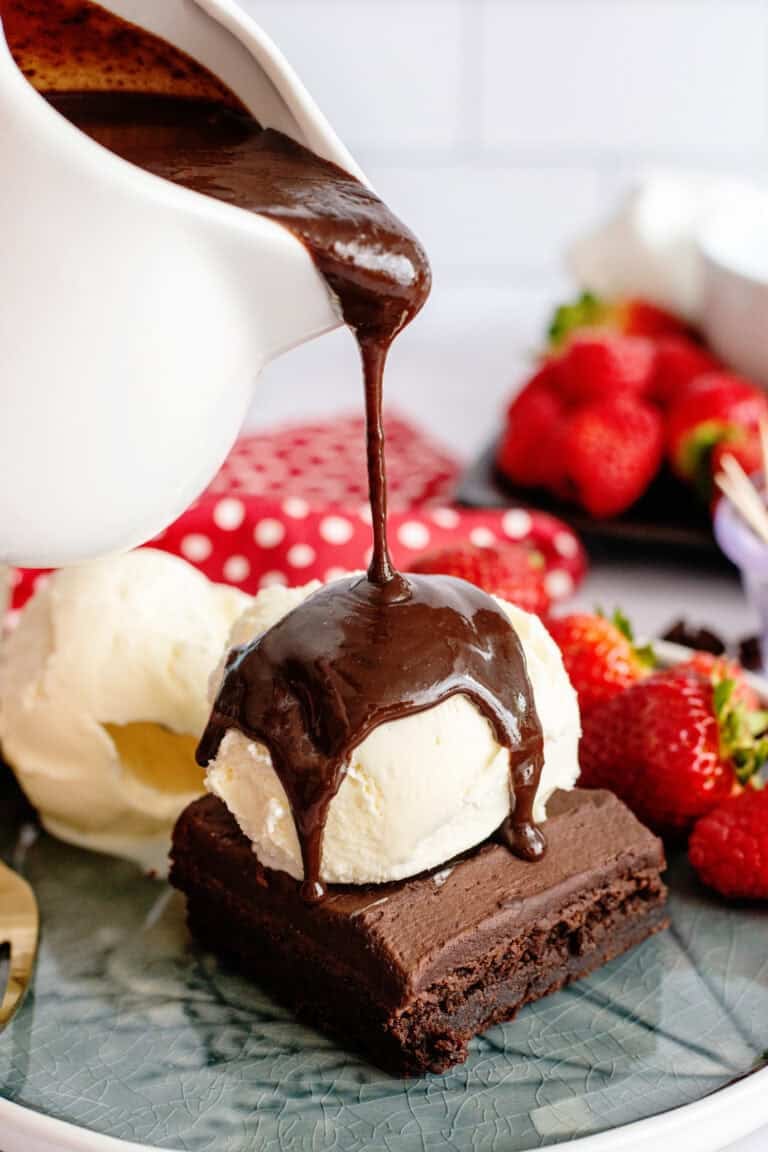 Hot fudge pouring from a pitcher onto a brownie topped with ice cream.