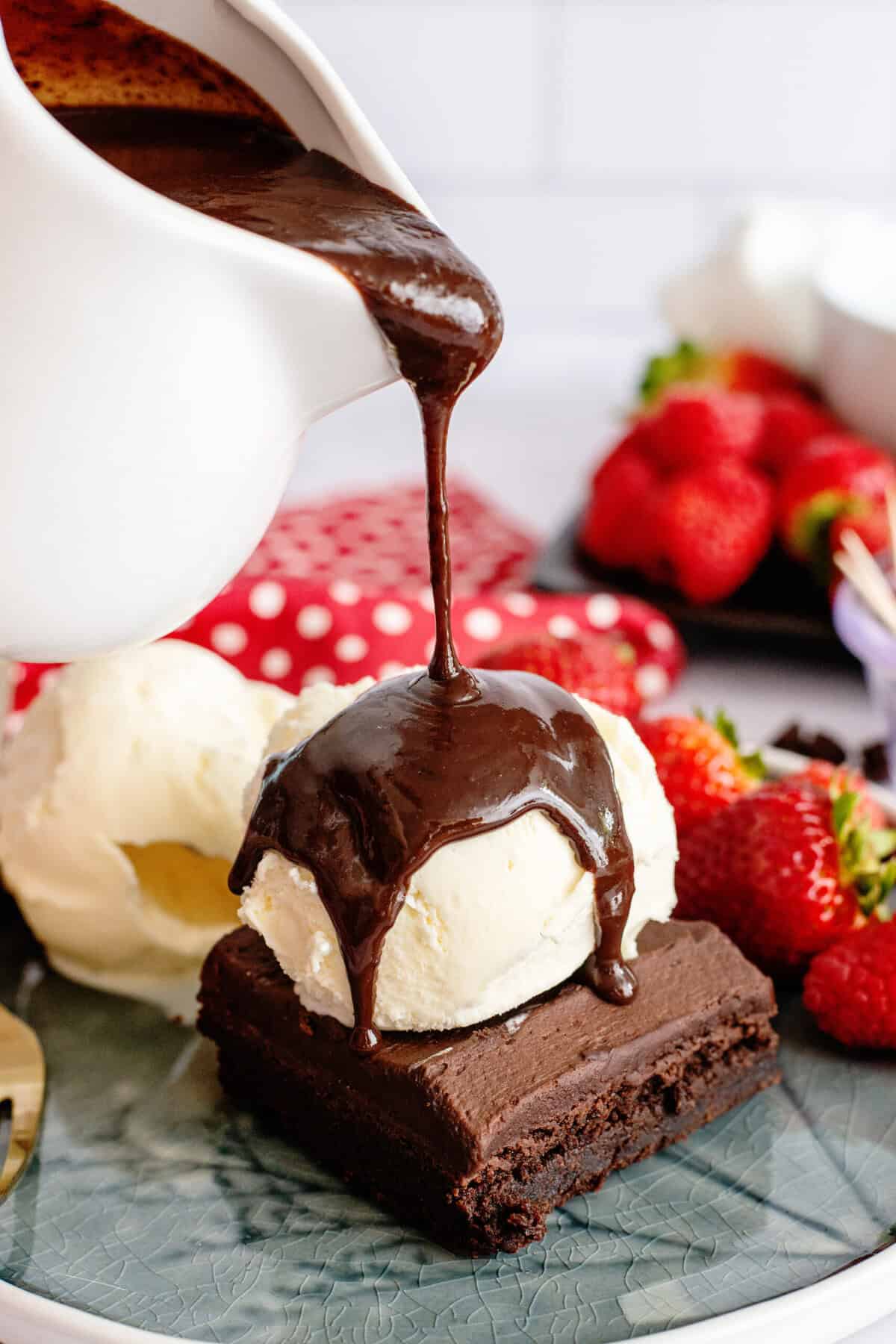 Hot fudge pouring from a pitcher onto a brownie topped with ice cream.