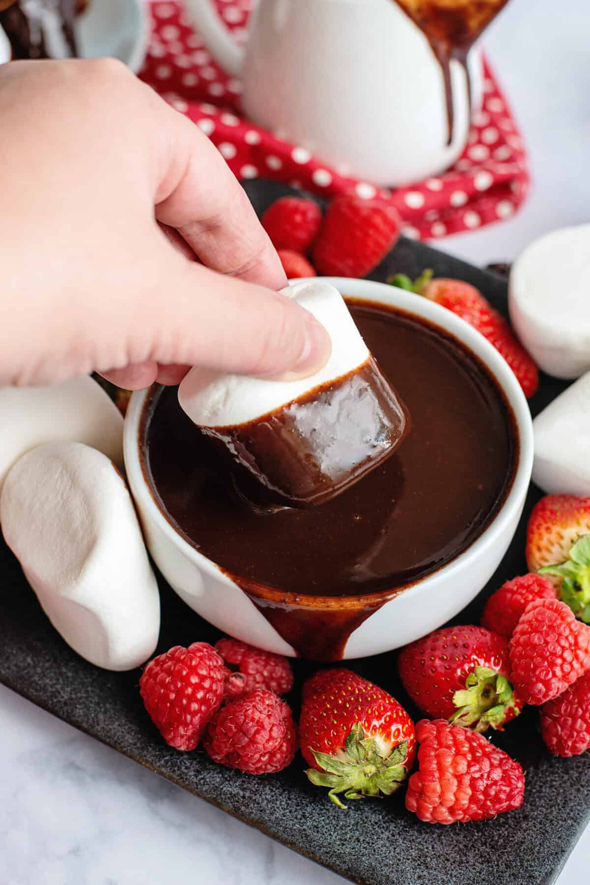 Dipping a marshmallow into a bowl of hot fudge surrounded by strawberries.