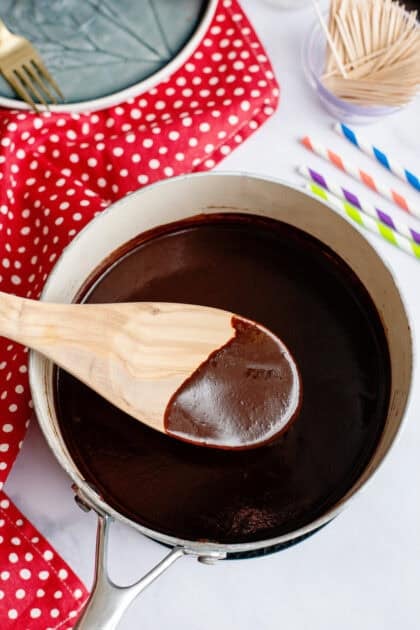 A wooden spoon dipped in hot fudge hovering over a saucepan.