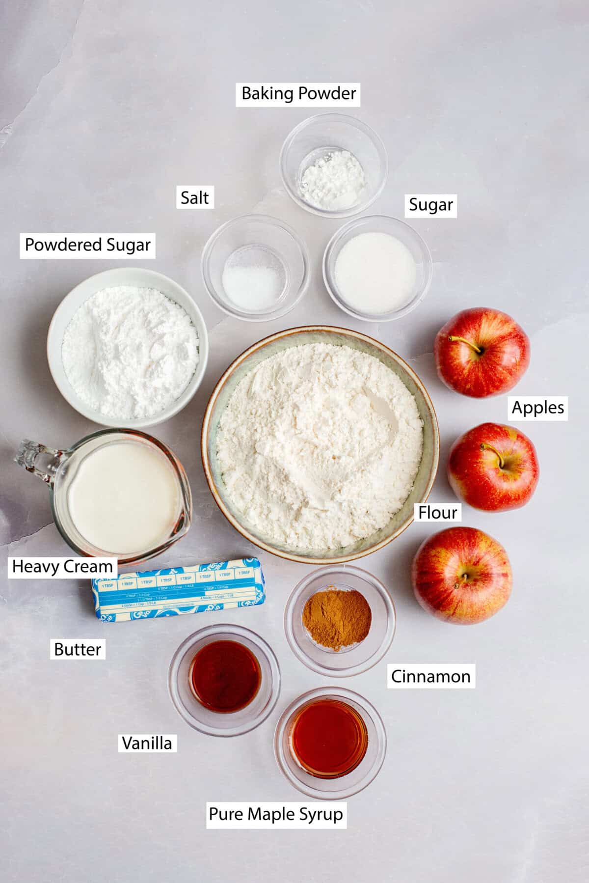 Ingredients for apple maple bakery scones, featuring all-purpose flour, sugar, baking powder, salt, butter, apples, cinnamon, heavy cream, vanilla, powdered sugar, maple syrup, and milk.