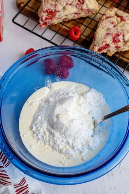 Mixing powdered sugar, vanilla extract, and milk in a glass blue mixing bowl.