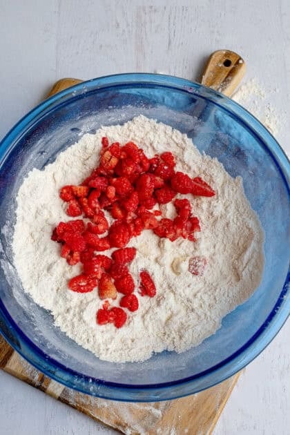 Adding raspberries to a glass blue mixing bowl of crumbly scone dough.