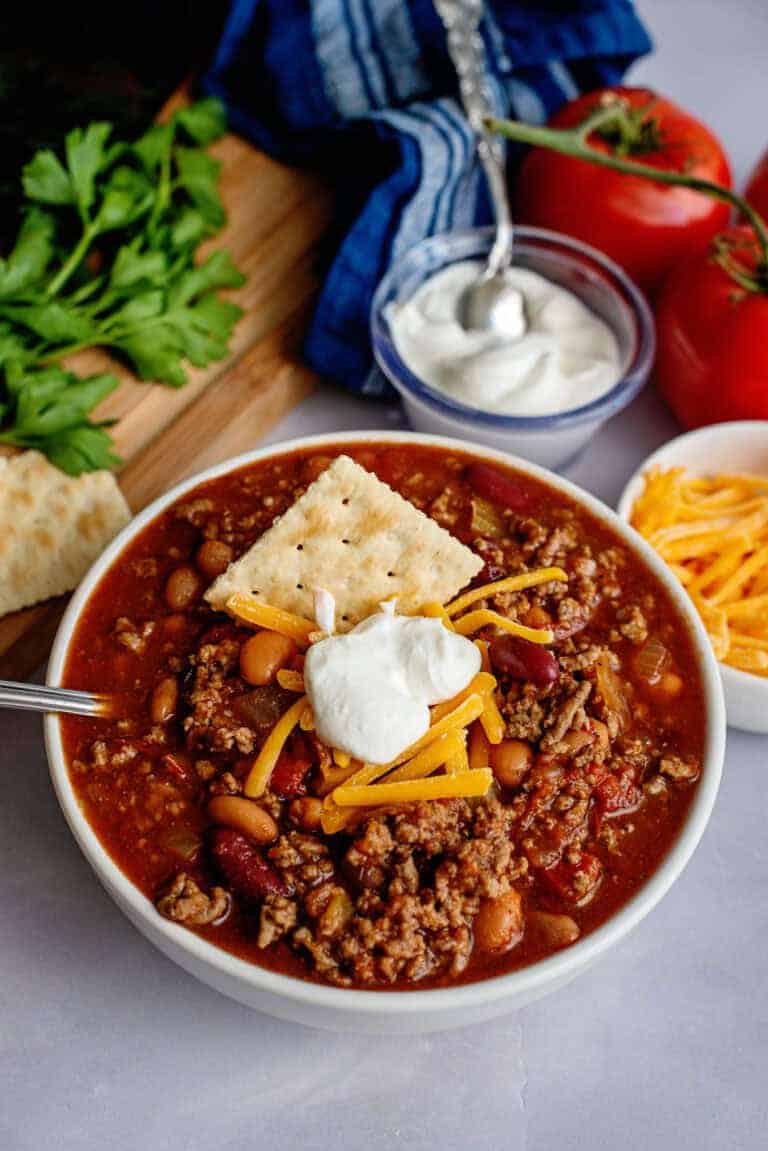 a bowl of hearty crockpot cincinnati chili with a dollop of sour cream, cheese, and a cracker on top