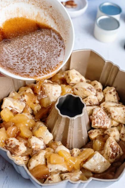 Pouring melted butter brown sugar mixture over apple pie filling coated cinnamon rolls in a bundt cake.