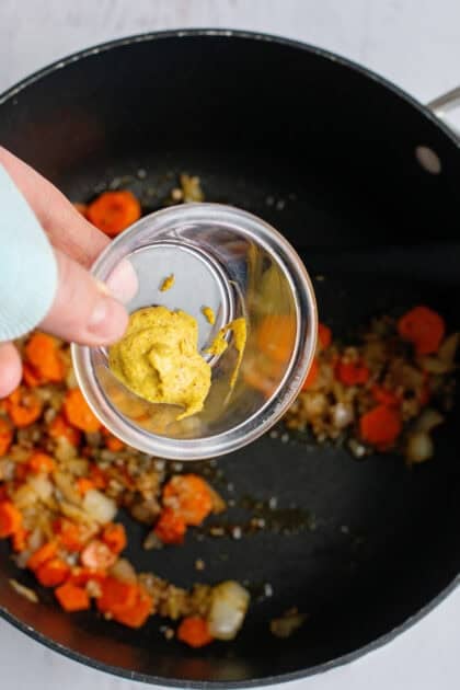 Adding mustard to a pot with carrots, onions, garlic and spices.