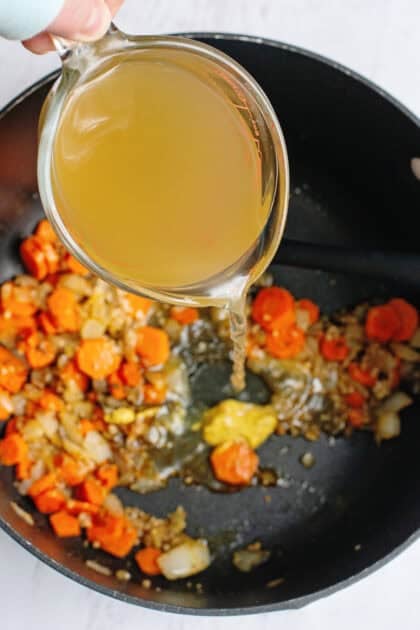 Adding broth to a pot with carrots, onions, garlic and spices.