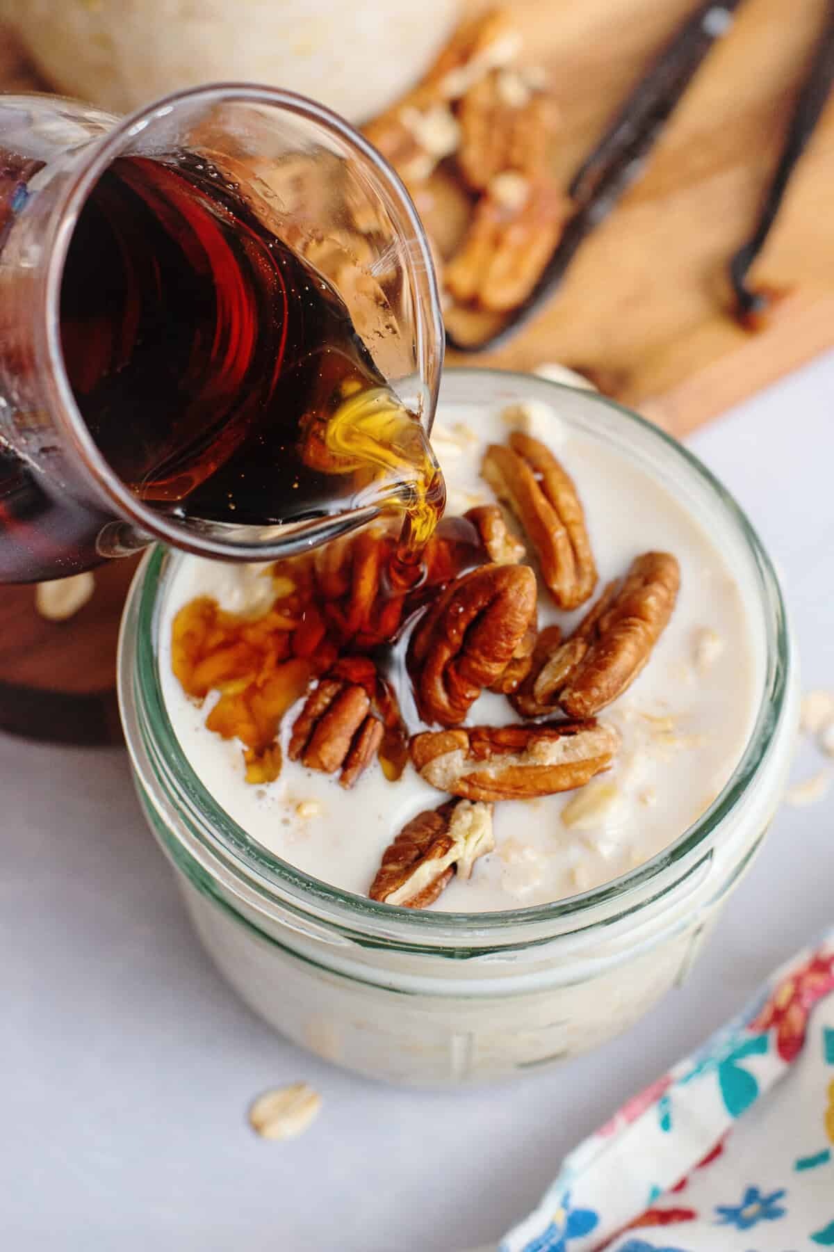 Pouring maple syrup into a jar of overnight oats with pecans.
