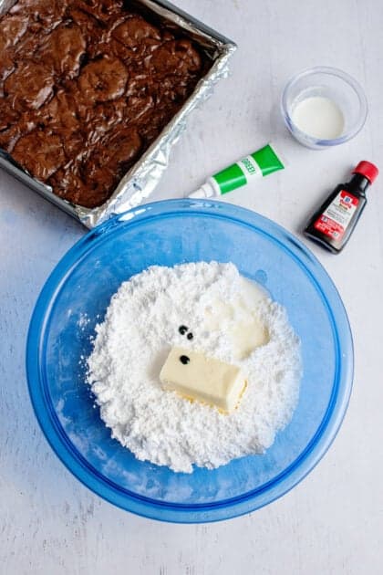 Butter, powdered sugar, heavy cream, and green food coloring in a glass blue mixing bowl.