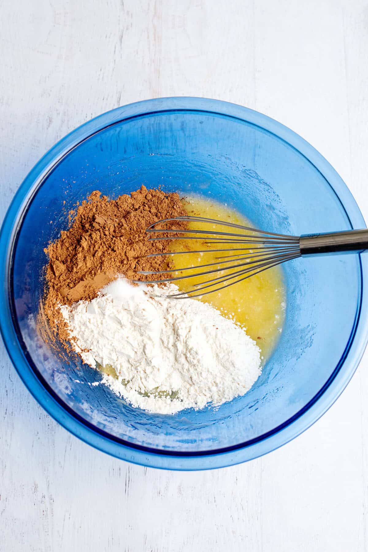Whisking cocoa powder, flour, and salt in a glass blue mixing bowl with whisked eggs.