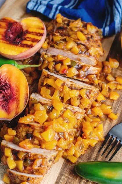 Grilled peach jalapeno glazed pork tenderloin sliced on a cutting board surrounded by fresh peaches and jalapenos.