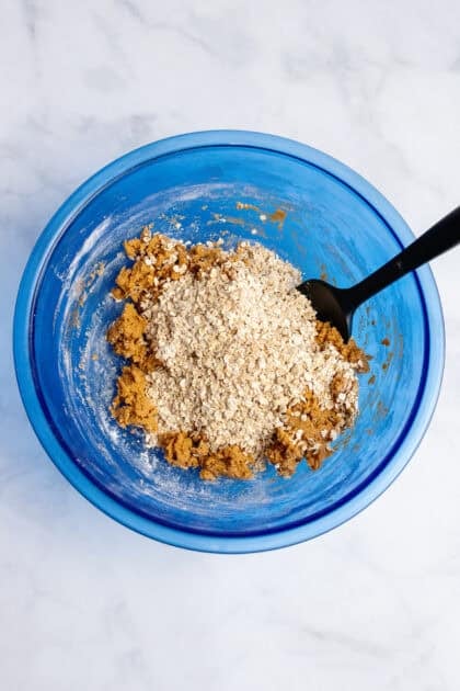 Adding oats to pumpkin cookie dough in glass blue mixing bowl.