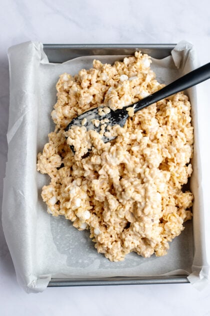 A black spatula spreading Rice Krispies into a parchment lined baking pan.